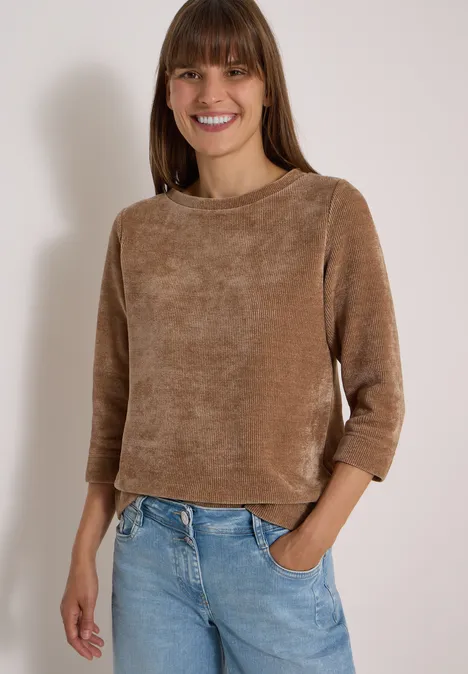 Een lachende vrouw in een bruine chenilletrui en lichtblauwe jeans, hand in zak, voor een lichte achtergrond.
