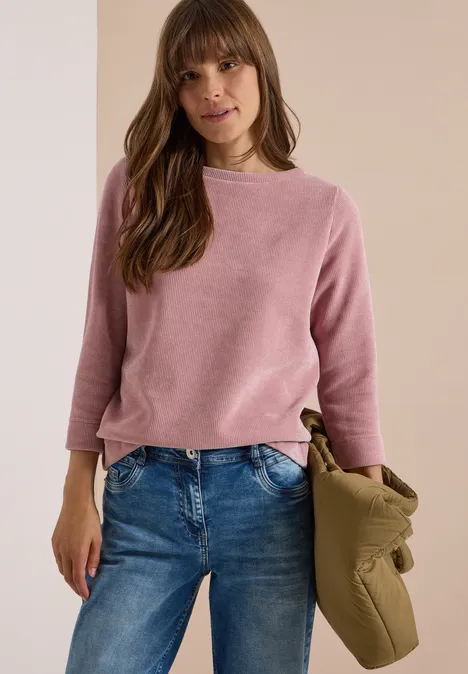 Frau in pinkem Pullover und Jeans, hält eine olivgrüne Jacke, stehend vor neutralem Hintergrund.