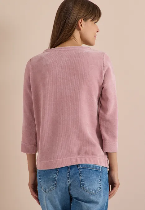 Person von hinten in rosafarbenem Kuschelpullover mit 3/4-Ärmeln und blauen Jeans vor beigem Hintergrund.