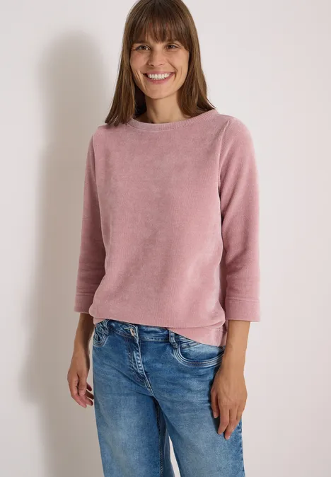 Frau in rosa Pullover und Jeans steht vor heller Wand, lächelnd in die Kamera blickend.