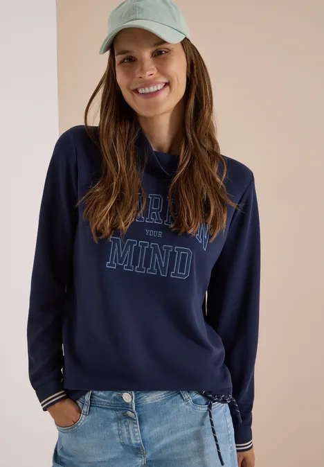 Koszulka z długim rękawem, stójką i napisem urban dark blue Młoda uśmiechnięta kobieta w dżinsach, granatowej bluzie 'OPEN MIND' i jasnozielonej czapce na jasnym tle.