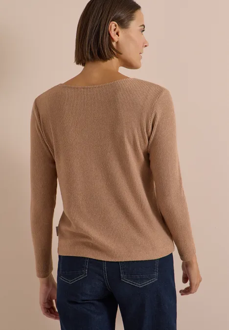 Frau mit kurzem Braunhaar steht mit dem Rücken zur Kamera, trägt beigefarbenen Pullover und dunkle Jeans.