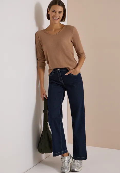 Frau in braunem Pullover und weiten Jeans lehnt lässig an der Wand und hält eine grüne Tasche.