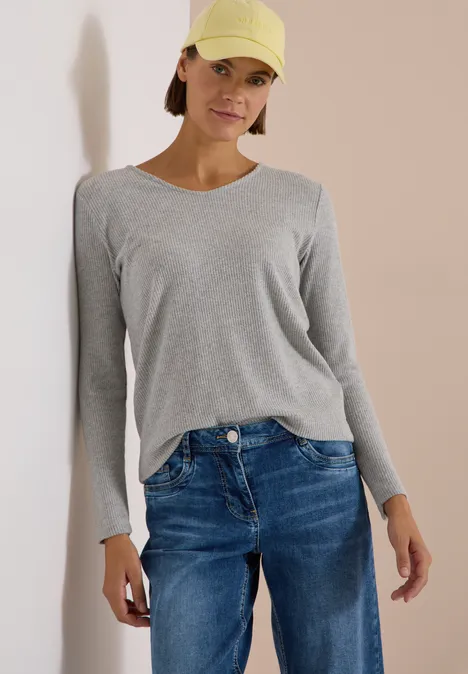 Frau mit grauem Langarmshirt, blauen Jeans und gelber Cap, stehend vor hellem Hintergrund.