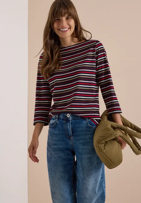 Femme en t-shirt à rayures et jean, tenant un manteau vert olive, souriante et détendue.