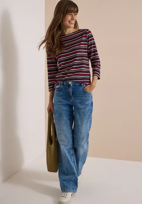 Jeune femme souriante en pull rayé, jean large et baskets blanches, appuyée contre un mur, avec un sac.