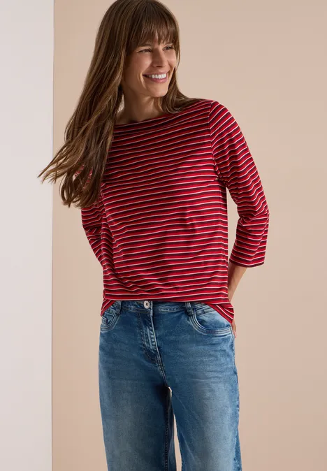 Femme souriante en t-shirt rayé rouge et noir et jean bleu, posant devant un fond neutre.