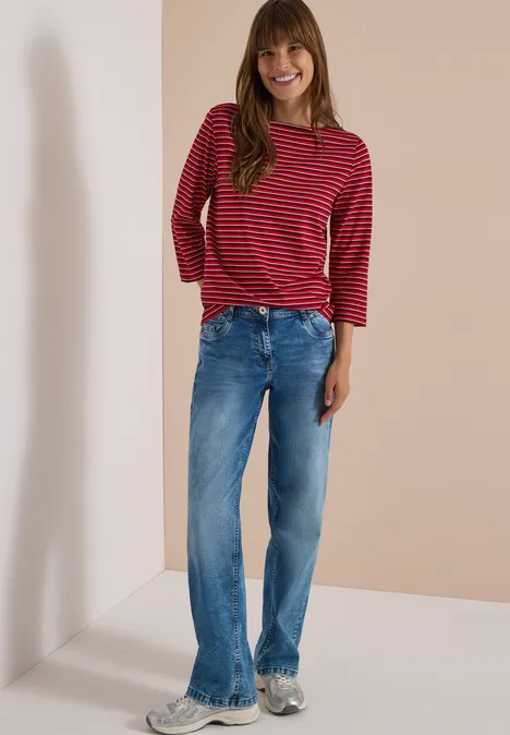 Jeune femme en jean et haut rouge à rayures blanches, souriante, se tenant près d’un angle, posture décontractée.