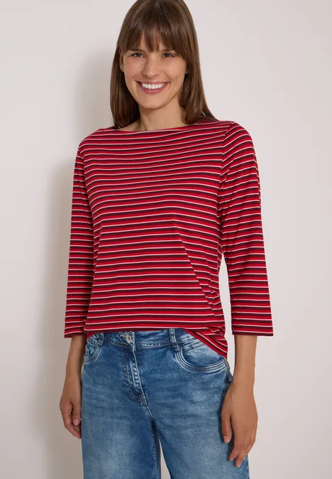 Femme souriante portant un haut à manches longues rayé rouge et bleu marine et un jean clair.