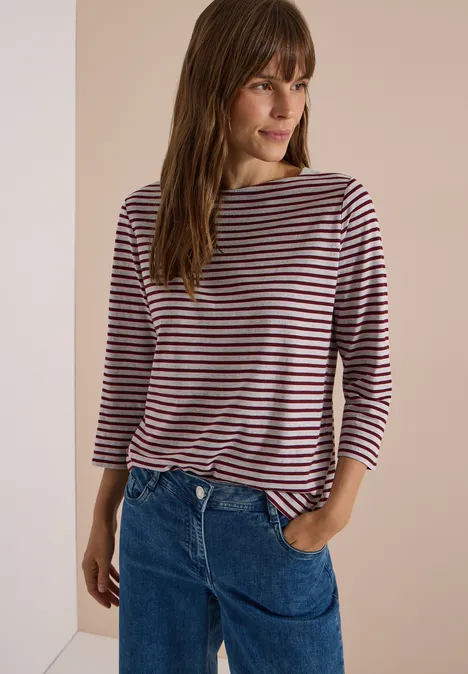 Frau mit braunem Haar in gestreiftem Langarmshirt und blauen Jeans vor neutralem Hintergrund.