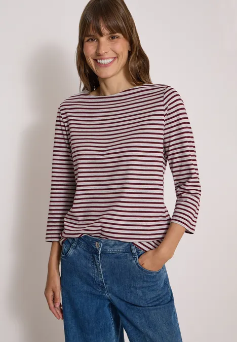 Frau lächelt in gestreiftem Langarmshirt und blauen Jeans, Hand in der Hosentasche vor neutralem Hintergrund.