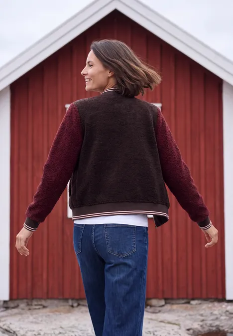 Vrouw met bruin haar in jeans en vest, armen wijd uitgestrekt voor een rode schuur.