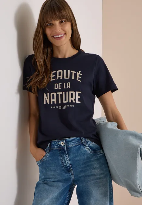 Une femme souriante porte un t-shirt bleu foncé « Beauté de la nature », un jean et tient une veste.