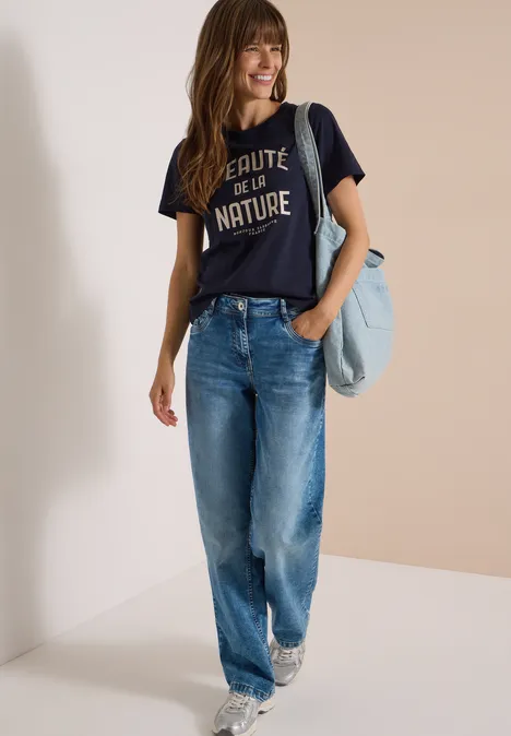 Une femme souriante en t-shirt sombre et jean décontracté porte un sac à l’épaule.