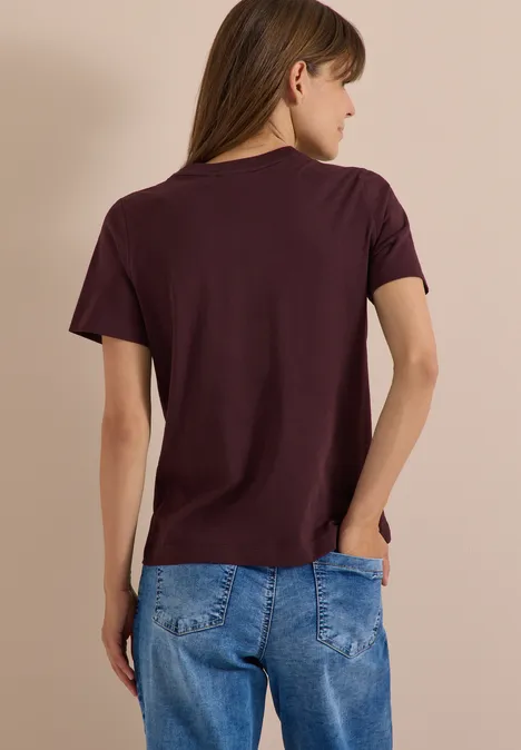 Une femme de dos portant un t-shirt bordeaux et un jean bleu devant un fond clair.
