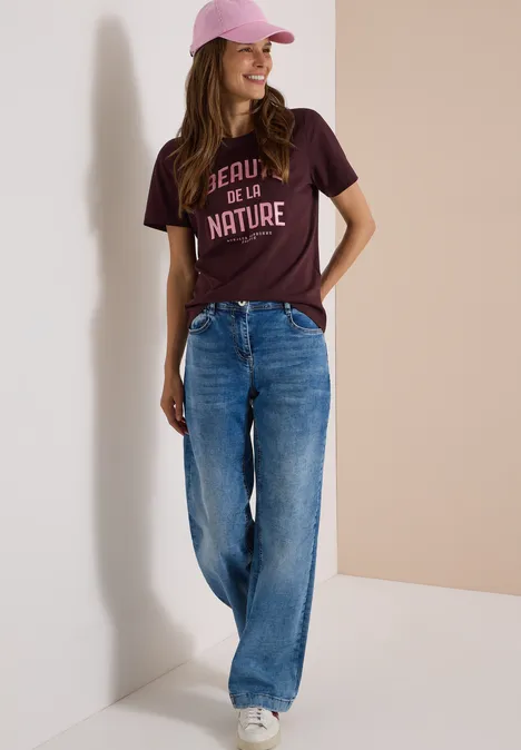 Femme souriante, t-shirt bordeaux imprimé, jean large et casquette rose sur fond clair
