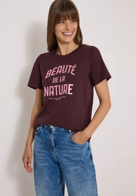 Femme portant un t-shirt bordeaux “Beauté de la nature” et un jean bleu décontracté sur fond blanc.