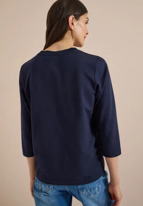 Tylne ujęcie kobiety w granatowym t-shircie z rękawem 3/4 i niebieskich dżinsach na beżowym tle.