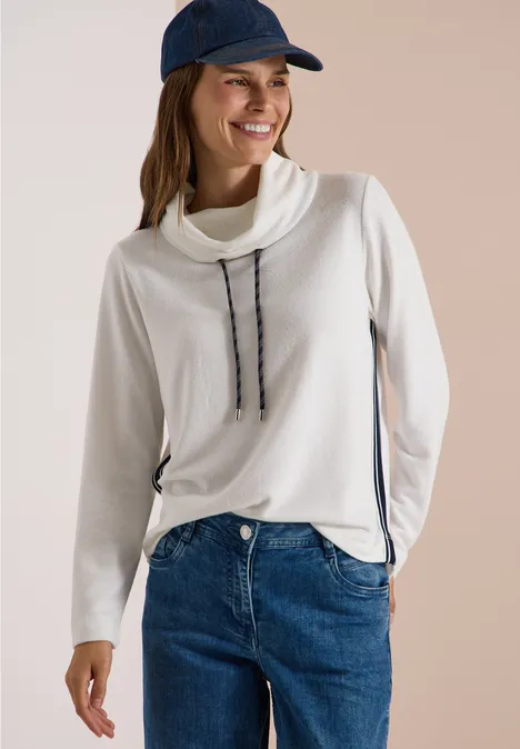 Zacht shirt met kraag en band vanilla white Lächelnde Frau im weißen Rollkragenpullover mit Kordeln, blauer Jeans und dunkler Baseballkappe.