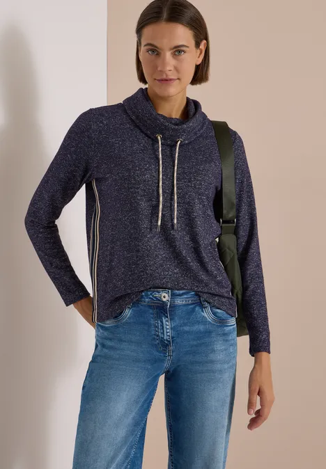 Femme en pull bleu marine col boule à cordons et jean, posant décontractée devant un mur beige.