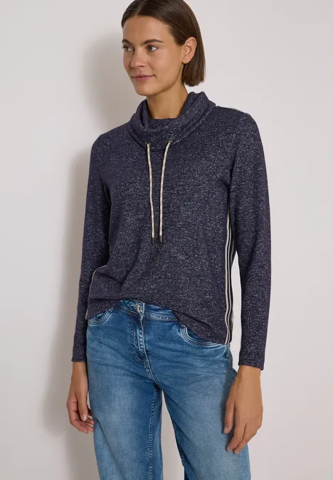 Femme portant un pull bleu foncé à cordon et un jean, debout devant un mur clair.