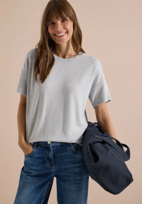 Eine lächelnde Frau in grauem T-Shirt und Jeans trägt eine dunkelblaue Tasche vor hellem Hintergrund.
