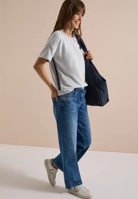 Frau in weißem T-Shirt und weiten Jeans trägt eine blaue Tasche und weiße Sneaker vor hellem Hintergrund