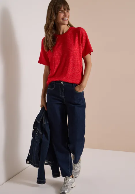 Frau in rotem T-Shirt, weiten Jeans und weißen Sneakers, hält eine Jeansjacke, steht vor hellem Hintergrund.