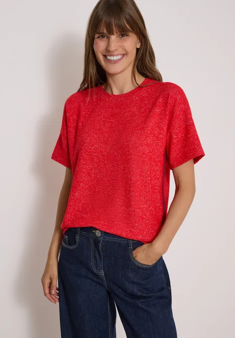 Lächelnde Frau in rotem Kurzarmshirt und blauer Jeans, eine Hand in der Hosentasche.