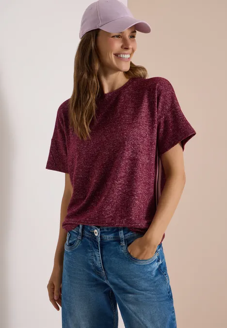 Frau trägt lila Cap, lockeres bordeauxfarbenes Shirt und Jeans, lächelt und hat Hände in den Taschen.