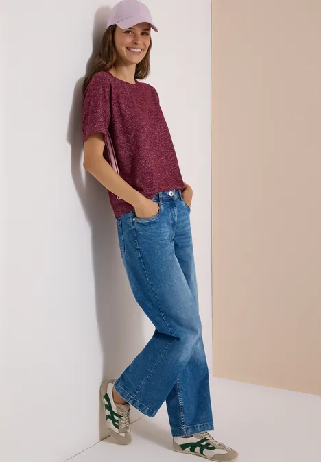 Frau in lockerem Freizeitoutfit (lila Shirt, Jeans, Cap, weiße Sneaker) lehnt lächelnd an heller Wand.
