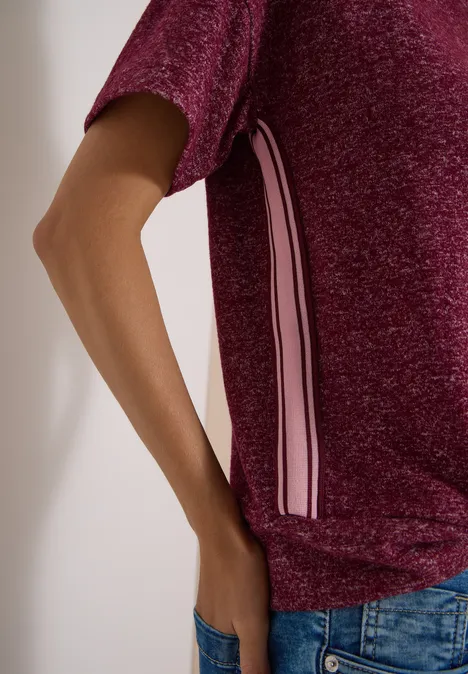 Person trägt glänzendes bordeauxfarbenes Shirt mit weißen und rosa Seitenstreifen, Hand in Jeans-Tasche.