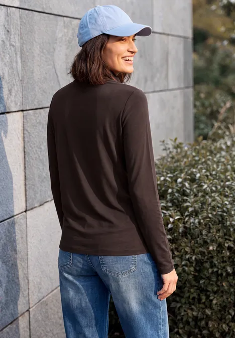 Frau im hellblauen Cap, schwarzem Langarmshirt und Jeans steht lächelnd an einer Mauer vor Büschen.