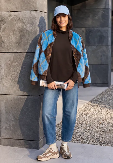 Frau mit Cap, blackem Shirt, blau gemusterter Jacke, Jeans und Sneakern lehnt an grauer Steinmauer