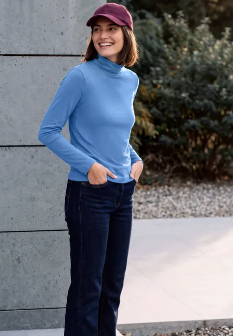 Frau mit roter Mütze, blauem Rollkragenpullover und Jeans steht lächelnd im Freien mit den Händen in den Taschen.