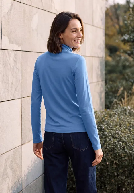 Frau in blauem Langarmshirt und Jeans lehnt an einer Steinwand und lächelt über die Schulter.