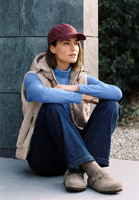 Eine junge Frau im lässigen Outfit sitzt auf dem Boden, lehnt an einer Steinmauer und schaut nach rechts.
