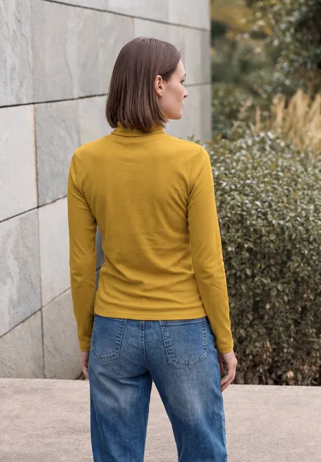 Langarm Shirt in Unifarbe mit Turtle Neck golden honey Frau in gelbem Langarmshirt und Jeans steht im Freien vor einer Steinwand und Büschen.