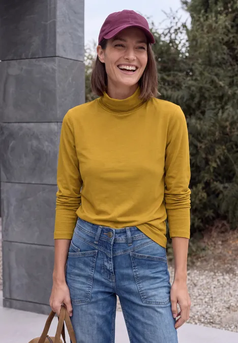 Langarm Shirt in Unifarbe mit Turtle Neck golden honey Eine lächelnde Frau in senfgelbem Rollkragenpullover und Jeans steht draußen und hält eine Tasche.