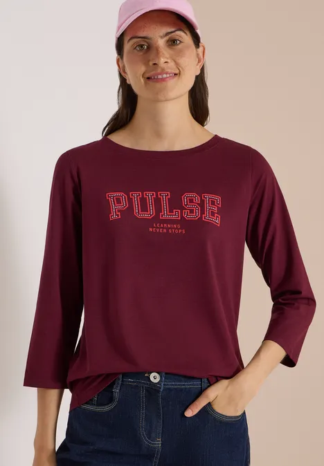 Femme en tee-shirt bordeaux à manches longues 'PULSE', jean et casquette rose, souriante.