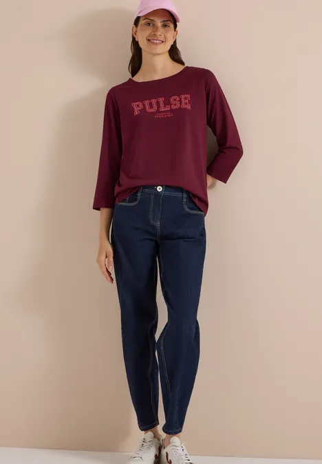 Femme avec casquette rose, t-shirt Pulse bordeaux, jean bleu et baskets blanches devant un mur beige.