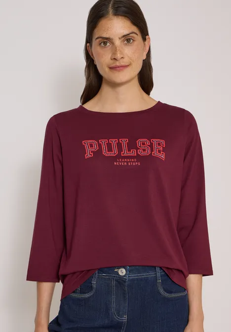 Femme portant un t-shirt bordeaux avec l'inscription 'PULSE' et un jean foncé.