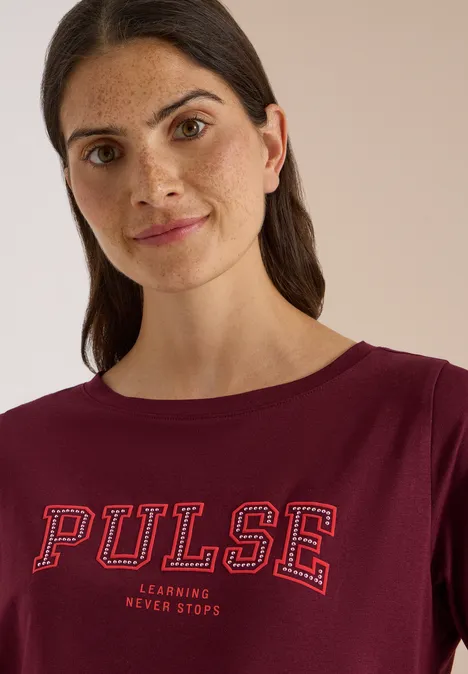 Une femme aux cheveux bruns porte un t-shirt bordeaux marqué 'PULSE' et regarde l'objectif avec un léger sourire.