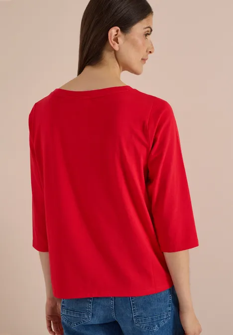 Femme vue de dos, porte un tee-shirt rouge à manches trois-quarts et un jean bleu, regard latéral.