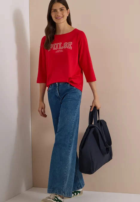 Jeune femme en t-shirt rouge 3/4 'POOLS', jean évasé bleu, souriante, sac bleu marine.