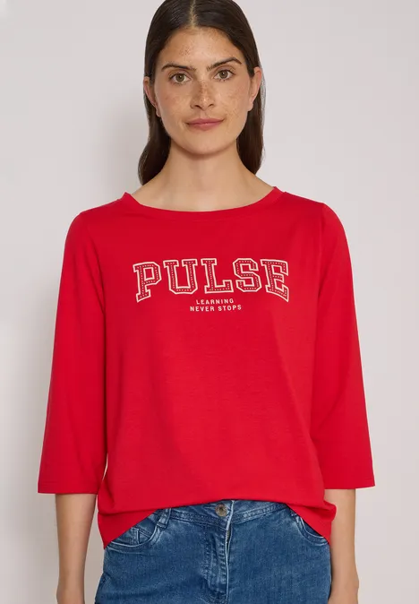 Jeune femme devant fond clair porte un t-shirt rouge 'PULSE' et un jean bleu.
