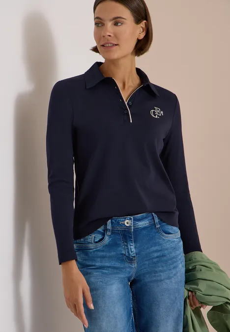 Frau in dunkelblauem Langarm-Poloshirt mit Reißverschluss und Jeans, hält eine grüne Jacke.