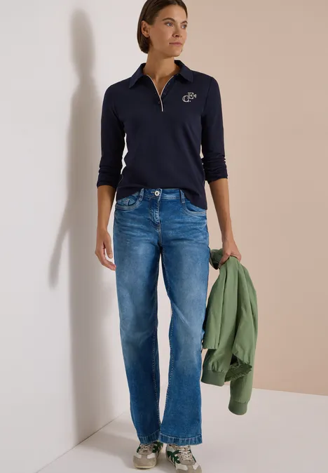 Eine Frau trägt ein navy Polo, blaue Jeans und hält eine grüne Jacke.