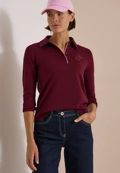 Koszulka Polo z długim rękawem i guzikami red wine Kobieta w bordowej koszulce polo z rękawami 3/4, niebieskich dżinsach i różowej czapce.