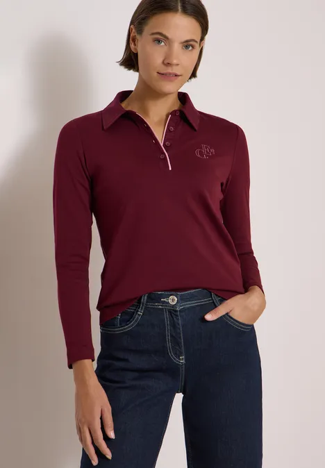 Koszulka Polo z długim rękawem i guzikami red wine Kobieta w bordowej bluzce polo z długim rękawem i niebieskich jeansach, swobodnie stojąca z ręką w kieszeni.